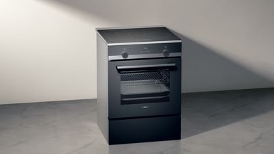 Moderne sort fritstående komfur med glaslåge og betjeningsknapper mod en minimalistisk lys væg og gulv. 