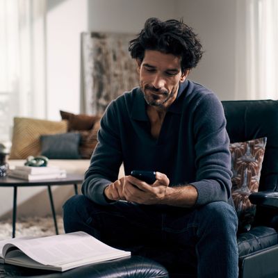 En mand sidder i en sofa og kigger på sin smartphone, omgivet af en hyggelig stue med naturligt lys og stilfuld indretning. 