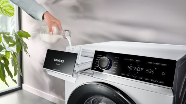 Máquina de lavar com exposição digital, detergente de derramamento de mão, planta de casa em fundo. 